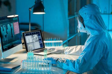 Side view of contemporary African American virologist in protective workwear sitting by desk in front of laptop and computer monitor