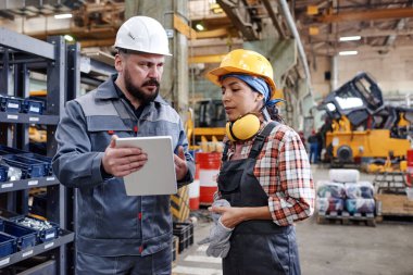 Bearded mature engineer with tablet making presentation of new technical project to young female colleague in factory