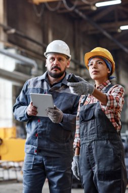 Engineers of large contemporary industrial plant looking at huge machine while female regulating its work and man using tablet