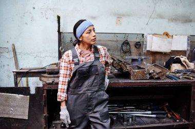 Young contemporary repairperson in coveralls standing by workbench with open toolkit and variety of worktools on shelves