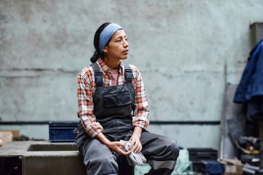 Young tired female engineer in coveralls and headband taking short rest after hard work in large warehouse of modern factory or plant