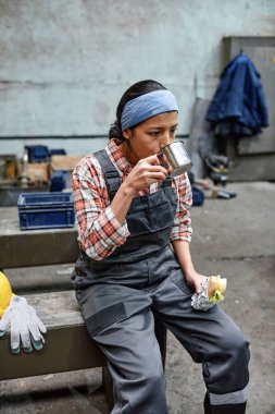 Young tired and hungry female engineer in coveralls having tea with sandwich for lunch at break after work in warehouse