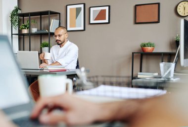 Serious young businessman with mug of tea looking through online data on laptop screen while analyzing it and having drink in office