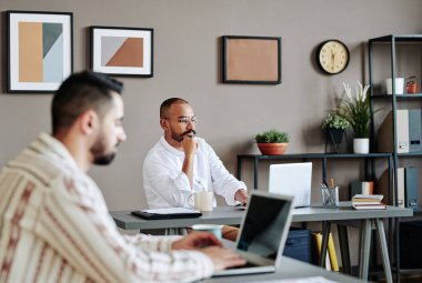 Young serious male ceo or analyst sitting by desk in front of colleague with laptop preparing report or presentation in office