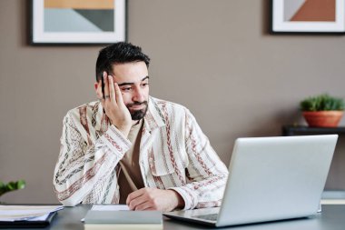 Young pensive businessman keeping hand on cheek during online communication with business partner at digital meeting
