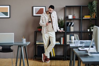 Contemporary young white collar worker in casualwear texting in smartphone while standing between desks with computers