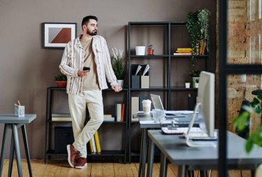Young businessman with smartphone looking through office window while standing against shelves with documents and green plants
