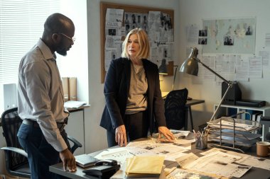 Two interracial FBI agents standing by desk with profiles of criminals and discussing their ideas about where to find them
