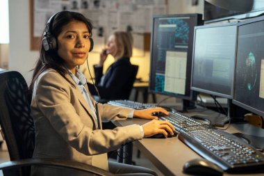 Young confident Hispanic female agent of secret service with headphones looking at camera while working in office