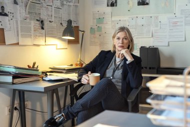 Serious mature female detective with cup of coffee sitting by workplace against walls with criminal profiles and other working documents