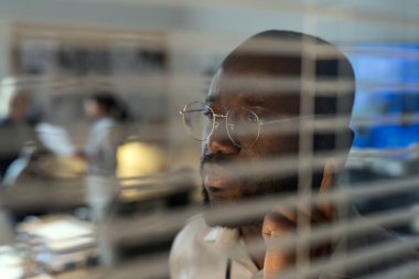 Serious African American FBI agent talking on the phone by venetian blinds while standing in office against two female colleagues