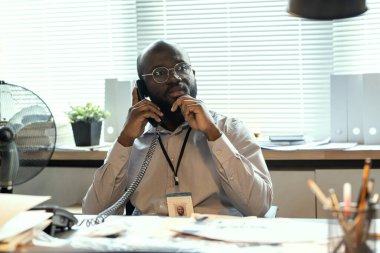 Contemporary male agent of FBI thinking of something while holding phone receiver by ear and talking to colleague on assignment