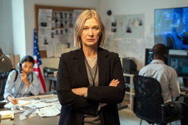 Serious mature leader of criminal department in formalwear standing in office against her two young interracial subordinates