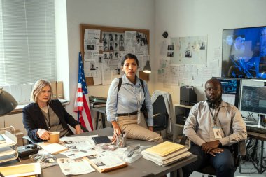 Group of three interracial agents of FBI or investigators sitting by workplace in office and looking at camera at meeting
