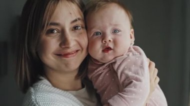 Medium close-up portrait of young brown-haired Biracial woman standing at home, holding baby girl and looking on camera at daytime