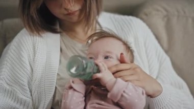 Tilting up of young short-haired Biracial woman sitting on couch at home, holding baby girl who drinking water from feeding bottle at daytime