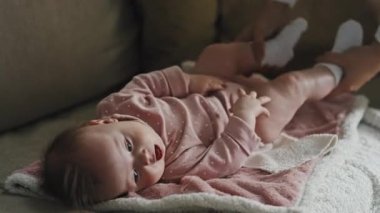 Locked-down of adorable Biracial baby girl in pink onesie lying on back on couch, hands of cropped person moving legs of infant