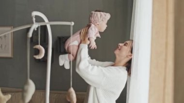 Medium of young short-haired mother holding adorable Biracial infant in pink hat at home in afternoon, admiring and lifting up baby