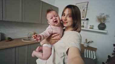 Handheld POV of young short-haired mother holding crying Biracial infant in kitchen at daytime, talking and looking on camera