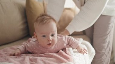 Medium long of cute blue-eyed Biracial baby girl lying on stomach on couch, cropped parent standing and watching her