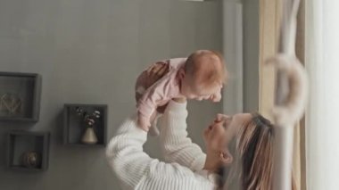 Side-view of cheerful young Biracial woman lifting up her cute baby girl in pink onesie, moving to sides, talking and smiling in living room at daytime
