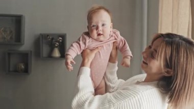 Side-view of cheerful young Biracial woman holding her cute baby girl in pink onesie in hands, lifting her up, talking and smiling in living room at daytime