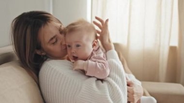 Side view of loving young Biracial woman sitting on couch, holding and kissing her cute baby daughter in pink onesie, talking and smiling in living room at daytime