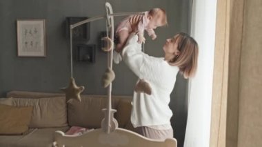 Medium side view of joyous young Biracial woman lifting up her cute baby daughter in pink onesie, talking and smiling at home in afternoon