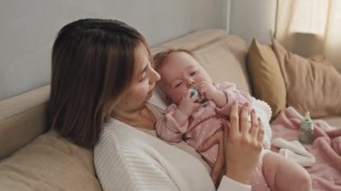 Blue-eyed Biracial infant in baby pink onesie putting pacifier in her mouth, resting in arms of her young mother on couch in afternoon