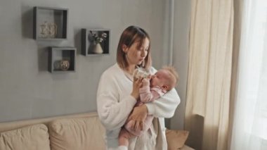 Medium of young Biracial parent standing in living room, holding crying blue-eyed daughter, giving her water in feeding bottle, rocking infant at daytime