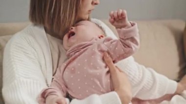 Medium of young Biracial parent sitting on couch, holding crying blue-eyed daughter, giving her water in feeding bottle at daytime