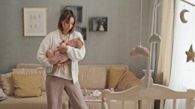 Medium long of young Biracial woman standing in living room, holding adorable blue-eyed daughter, rocking her at daytime