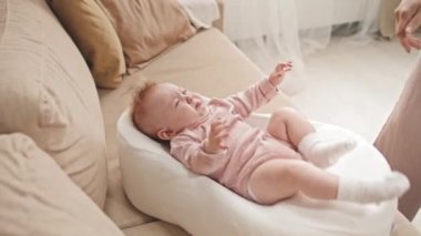 Blue-eyed crying Biracial infant in pink onesie lying in baby nest on couch at home, cropped caretaker giving her pacifier