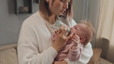 Tilting up of young Biracial parent standing in living room, holding crying blue-eyed daughter, giving her water in feeding bottle, rocking infant at daytime