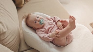 Top view of cute blue-eyed Biracial infant in pink onesie with pacifier lying in baby nest on couch at home, looking on camera