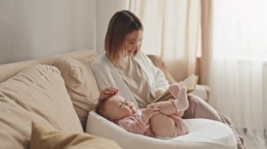 Medium long of brown-haired young woman sitting on couch in living room at daytime, reading book to cute daughter in pink onesie who lying in baby nest