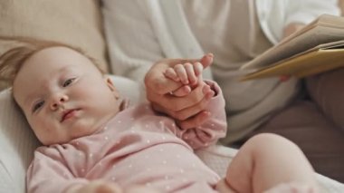 Medium long of adorable blue-eyed Biracial female child in pink onesie lying in baby nest on couch at home, holding hand of cropped parent, who reading book