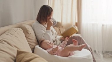 Medium long of brown-haired young woman sitting on couch in living room at daytime, reading book and drinking coffee, cute daughter in pink onesie lying in baby nest