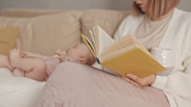 Midsection of cropped female babysitter sitting on couch in living room at daytime, reading book and drinking coffee, blurred infant lying in baby nest