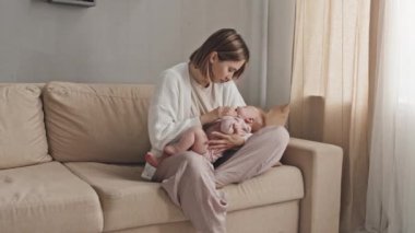 Young short-haired Biracial woman sitting on couch in living room at daytime, holding her crying baby daughter, giving her pacifier