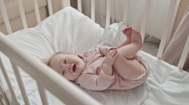 Top down of happy newborn girl in pink onesie lying in crib, smiling and looking away at daytime