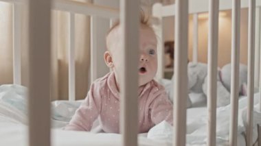 Low angle of cute blue-eyed baby girl with messy hair lying on stomach in crib, smiling at daytime