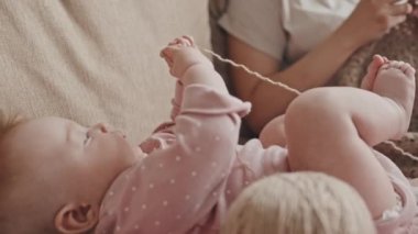 Low angle of unrecognizable mother sitting on couch, knitting, cute blue-eyed baby girl lying nearby, moving and playing with yarn in afternoon
