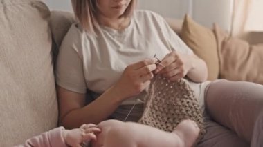 Tilting up of brown-haired Biracial woman in loungewear sitting on sofa at home, knitting while babysitting baby daughter lying nearby at daytime