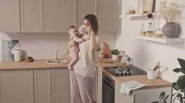 Medium long of young Biracial woman in loungewear holding cute baby girl, standing in kitchen at daytime, drinking coffee