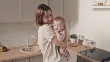 Medium of young short-haired Biracial woman holding blue-eyed baby girl, standing in kitchen at daytime