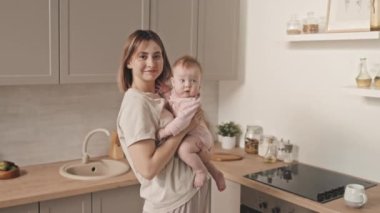 Medium portrait of young short-haired Biracial woman holding blue-eyed baby girl, standing in kitchen at daytime, looking and smiling on camera