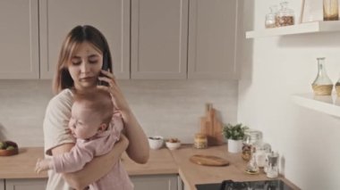 Medium of busy young Biracial woman holding beautiful baby daughter, talking on phone in kitchen in afternoon