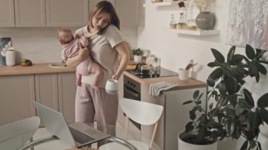 Medium long of busy young Biracial woman holding beautiful baby daughter, talking on phone, taking mug with coffee, using laptop computer in kitchen in afternoon