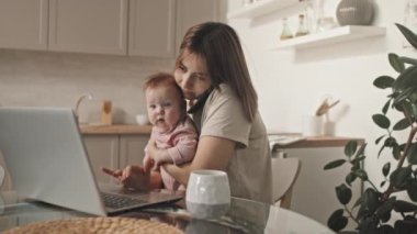 Medium of brown-haired young Biracial woman holding cute baby daughter, talking on phone, sitting at table, using laptop computer in kitchen in afternoon